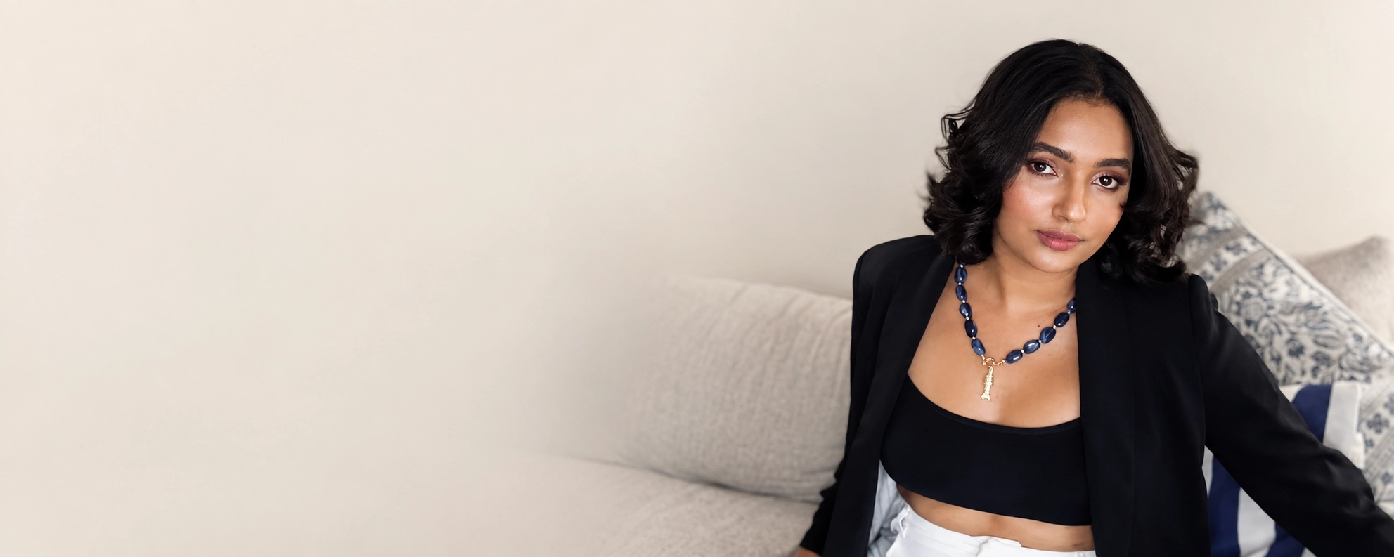 Karen Voguedo sentada en un sofá claro, con blazer negro y collar de piedras azules.